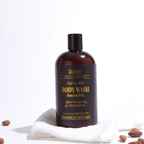 A bottle of Chocolate Bliss Body Wash with a dark label, resting on a white surface with a towel and some cocoa beans scattered around.